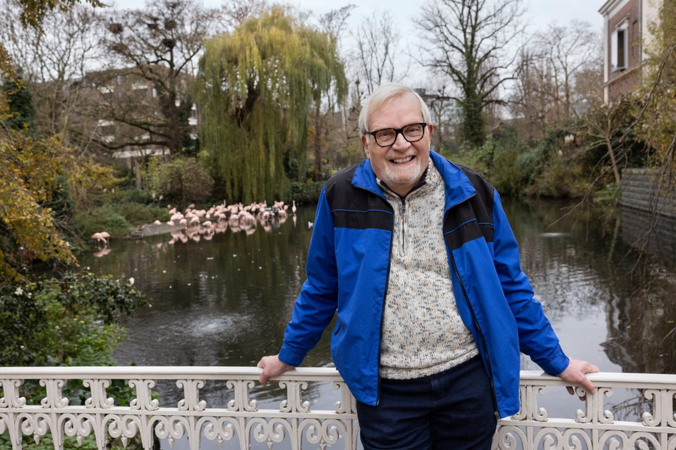 Man staat lachend leunend tegen hekje in Artis met flamingo's op de achtergrond